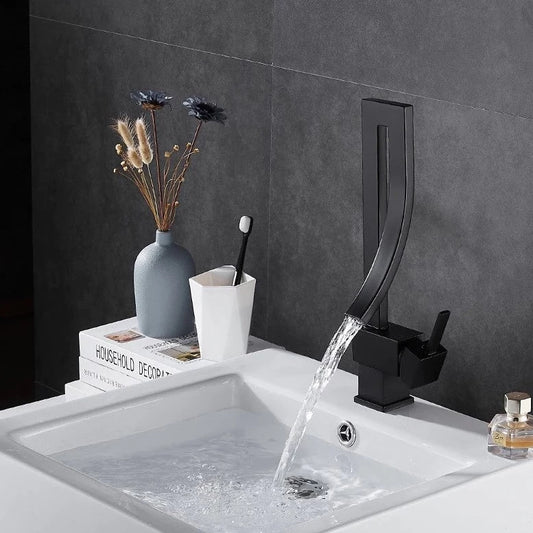 A modern, sleek Deck Mounted Brass Bathroom Basin Faucet from Bacala, featuring a curved design in black, is dispensing water into a white sink. In the background, there's a vase with dried flowers and a toothbrush holder containing a black and white toothbrush. The wall appears to be tiled in a dark color, echoing the elegance of a premium shower head.