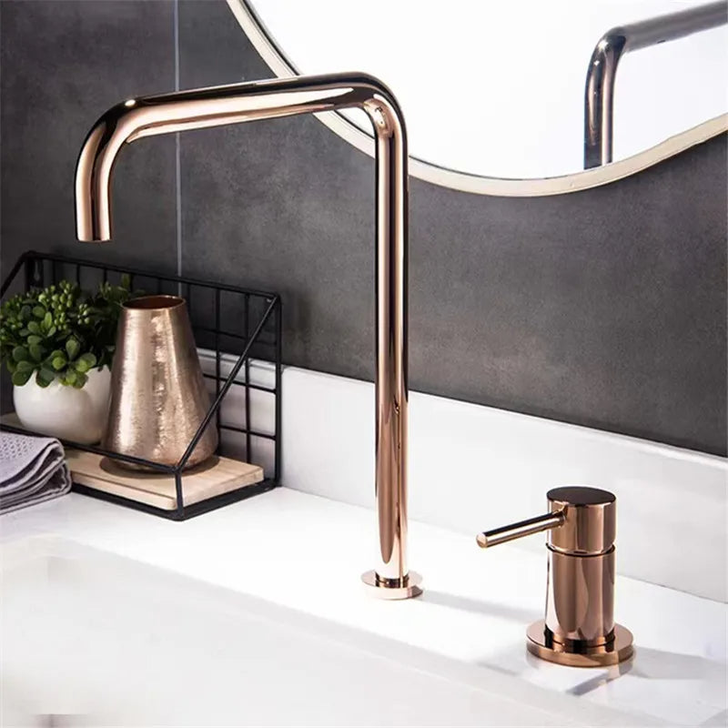 A modern bathroom sink setup featuring a rectangular white vessel sink with water flowing from AllFixture's sleek, curved Single Handle Rose Gold Bathroom Sink Faucet. Next to it is a matching rose gold soap dispenser. Complementing the scene is a grey tiled wall and a small plant adding a touch of green, reminiscent of chic kitchen faucet designs.