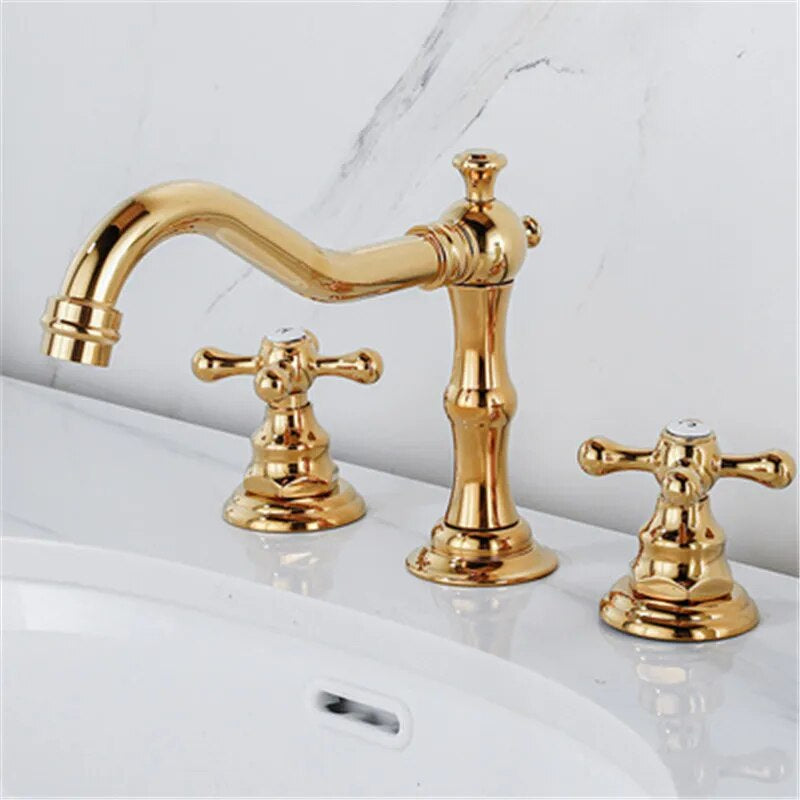 A polished gold Dual Handle Solid Brass Deck Mounted Bathroom Faucet from Tiqui set on a white countertop. The set features a curved spout with elegant detailing and two cross-handle knobs for hot and cold water control. The background is white with gray veining, resembling marble—truly an exquisite choice among bathroom faucets.