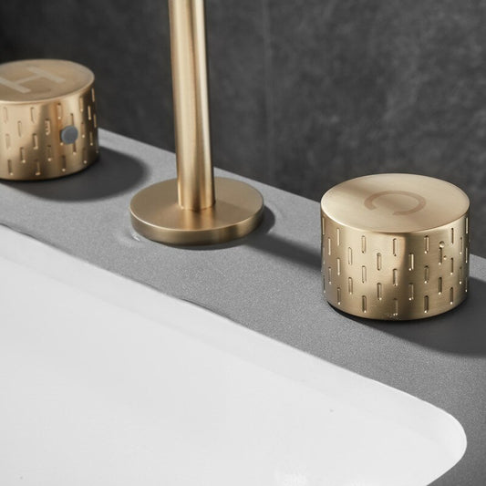Close-up of the Mike Jake Windsor Widespread Brass Faucet on a smooth gray countertop, featuring gold-colored handles, engraved symbols, a textured pattern, and durable ceramic disc valve technology.