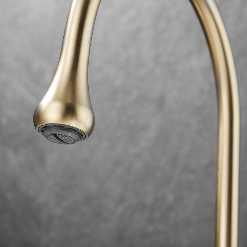 An elegant Tiqui™ Solid Brass Double Handle Bathroom Sink Faucet by Mike Jake with a sleek, curved, gold finish is installed on a white countertop. Water is flowing from the spout into the sink below. Two matching, textured gold handles are placed on either side of the faucet. The background features dark gray tile.