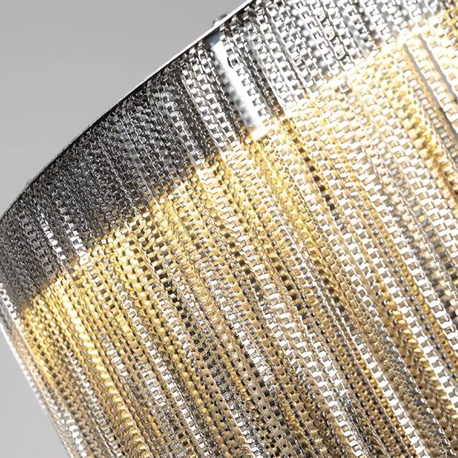Close-up view of a modern, textured, stainless steel lamp shade with warm LED lighting shining through the interwoven strips, creating a shimmering effect. The image captures the intricate pattern and reflective surface of the Morsale.com Cadena Steel Tassel Light Fixture.