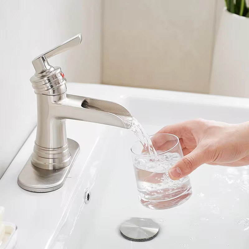 An Allfixture Single Handle Waterfall Bathroom Faucet, with its sleek modern design and stainless steel finish, is positioned over a white sink. Water cascades elegantly from the faucet into a clear glass held by a hand. In the background, beige-tiled walls and a glimpse of a green plant add to the aesthetic appeal.