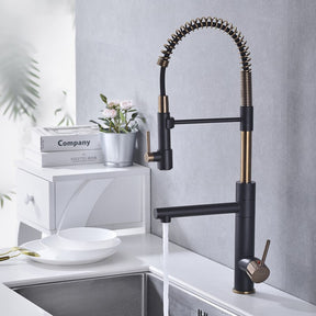 A modern kitchen sink with the stylish black and gold Tiqui™ Single Handle Pull Out Spout Kitchen Faucet by Smesiteli is seen. Water is flowing from the faucet into the sink. On the counter, there are white bowls, decorative items, a book titled "Company," and a potted plant.