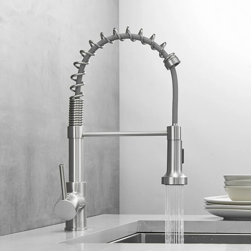 A person uses a modern Smesiteli Brushed Nickel Pull Out Spout Kitchen Faucet to rinse a white plate over a sink. The faucet, doubling as a chic bathroom sink fixture, boasts a high arc and pull-out spray head. The background features gray tiled walls, enhancing the sleek and contemporary look.