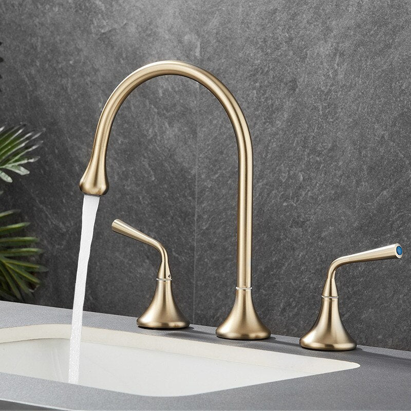 A sleek Tiqui™ Solid Brass Double Handle Bathroom Faucet by Mike Jake, featuring a high-arc spout, is mounted on a grey countertop. Water flows gracefully from the spout into a white sink. The background showcases a grey, textured wall and some green plant leaves peeking in from the left edge.