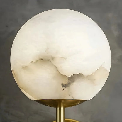 The Natural Marble Sphere Wall Sconce from Morsale.com, with unique sedimentary patterns in shades of white and gray, is mounted on a brass-colored stand, casting a warm glow against a soft gray background.