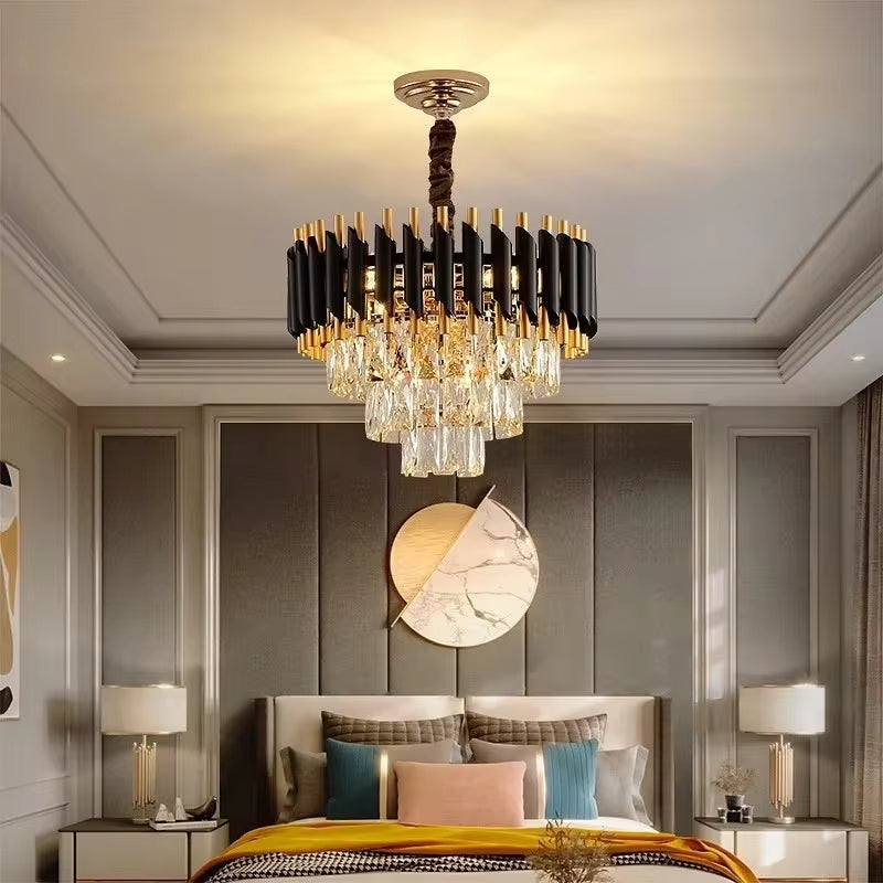 A contemporary bedroom features Morsale.com's Verano Crystal Chandelier with high-quality clear crystal accents suspended from the ceiling. The bed has a tufted, dark grey headboard and is decorated with various pillows. Above the headboard, there's a decorative circular wall art piece.