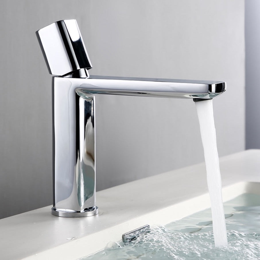 A sleek, modern Polished Chrome Bathroom Sink Faucet from Morsale.com, featuring a cylindrical base and a straight spout, pours a stream of water into a white sink. The background is a plain light gray wall.