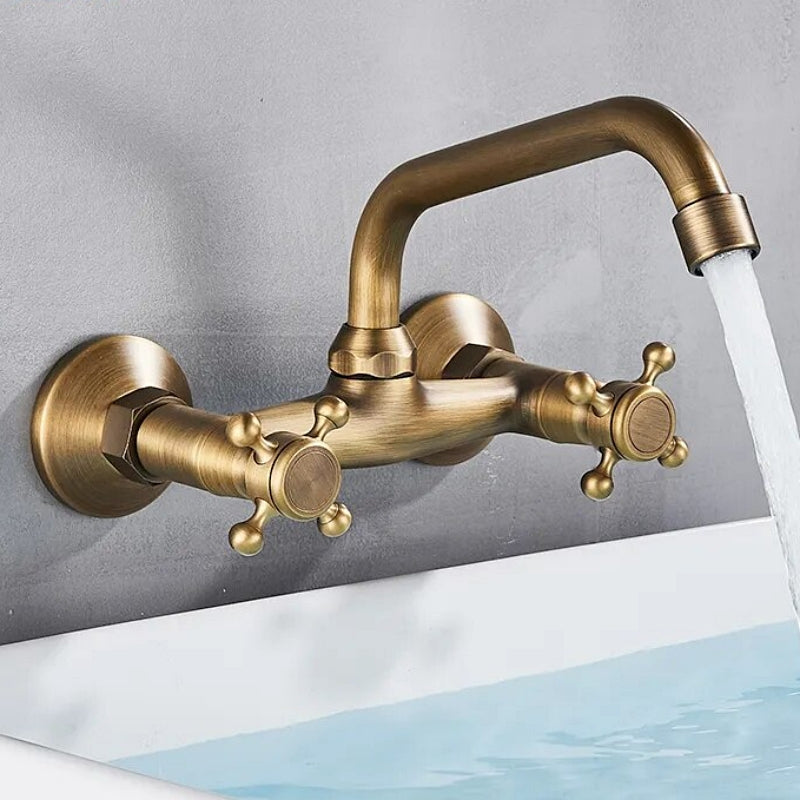 A Rozin Antique Brass Wall Mounted Bathroom Faucet with a vintage design is turned on, with water flowing into a sink or bathtub. These bathroom faucets feature two cross handles for hot and cold water, and the overall appearance gives a classic, antique feel.