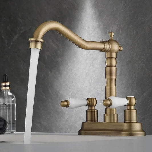 The "Antique Brass Bathroom Faucet - Timeless Elegance for Your Sink" by Allfixture stands over the sink, water flowing gracefully from its dual handle design with white handles. The background features a dark, marble-like surface. A soap dispenser is visible in the left corner of the image, complementing the premium brass construction.