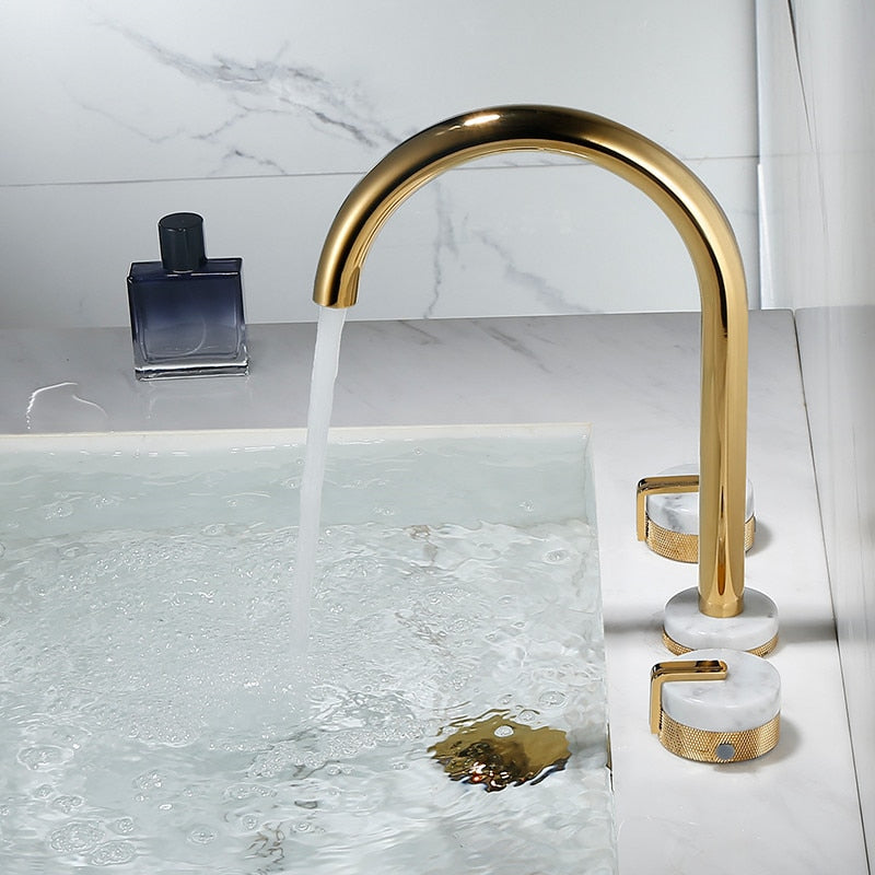 A modern bathroom sink features the Tiqui™ Solid Brass & Marble Bathroom Sink Faucet, with water gracefully flowing into a pristine white basin. Nearby, gold and marble handles enhance the ambiance, complemented by a dark blue bottle positioned on the elegant marble countertop. The background showcases a stunning marble-patterned wall.