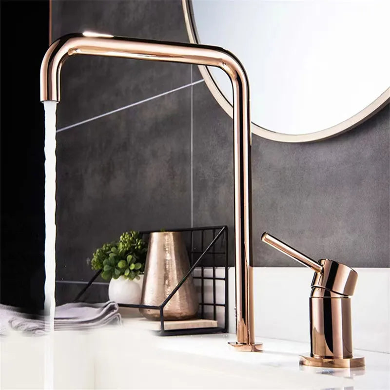 A modern bathroom sink setup featuring a rectangular white vessel sink with water flowing from AllFixture's sleek, curved Single Handle Rose Gold Bathroom Sink Faucet. Next to it is a matching rose gold soap dispenser. Complementing the scene is a grey tiled wall and a small plant adding a touch of green, reminiscent of chic kitchen faucet designs.