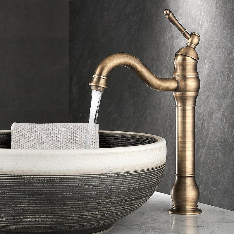 A Tiqui Deck Mounted Single Handle Bathroom Faucet is dispensing water into a decorative ceramic sink. A textured cloth is partially submerged in the water, draped over the edge of the sink. The background features a dark, textured wall surface.