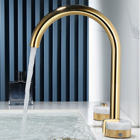 A modern bathroom sink features the Tiqui™ Solid Brass & Marble Bathroom Sink Faucet, with water gracefully flowing into a pristine white basin. Nearby, gold and marble handles enhance the ambiance, complemented by a dark blue bottle positioned on the elegant marble countertop. The background showcases a stunning marble-patterned wall.