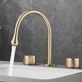 An elegant Tiqui™ Solid Brass Double Handle Bathroom Sink Faucet by Mike Jake with a sleek, curved, gold finish is installed on a white countertop. Water is flowing from the spout into the sink below. Two matching, textured gold handles are placed on either side of the faucet. The background features dark gray tile.