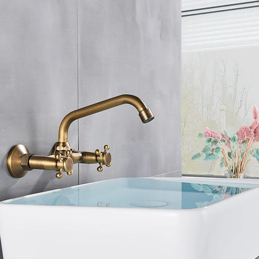 The Rozin Maison Brass Wall Mounted Bathroom Faucet in antique brass is installed above a modern white sink with water. A window with blinds and a vase of colorful flowers complete the serene bathroom scene.