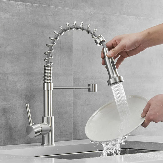 A person uses a modern Smesiteli Brushed Nickel Pull Out Spout Kitchen Faucet to rinse a white plate over a sink. The faucet, doubling as a chic bathroom sink fixture, boasts a high arc and pull-out spray head. The background features gray tiled walls, enhancing the sleek and contemporary look.