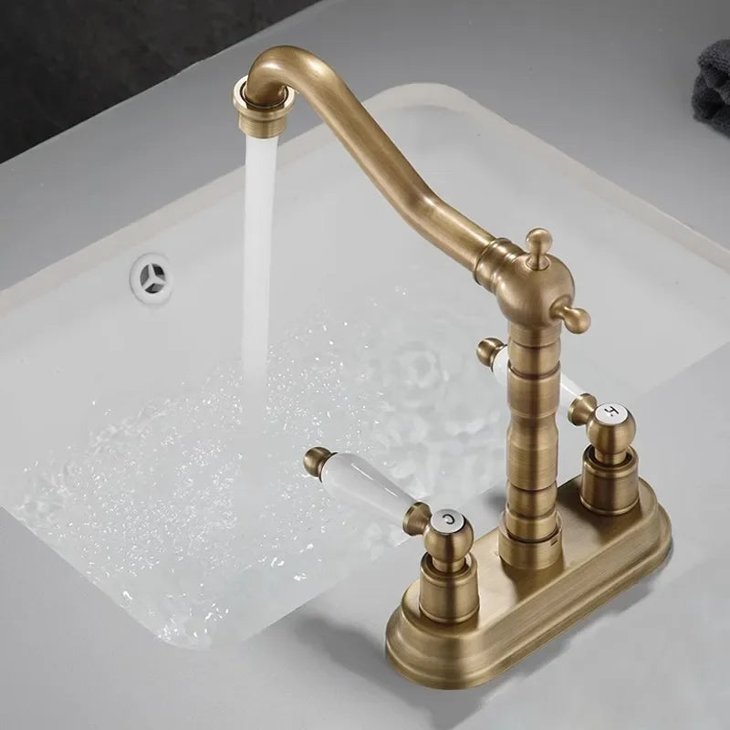 The "Antique Brass Bathroom Faucet - Timeless Elegance for Your Sink" by Allfixture stands over the sink, water flowing gracefully from its dual handle design with white handles. The background features a dark, marble-like surface. A soap dispenser is visible in the left corner of the image, complementing the premium brass construction.