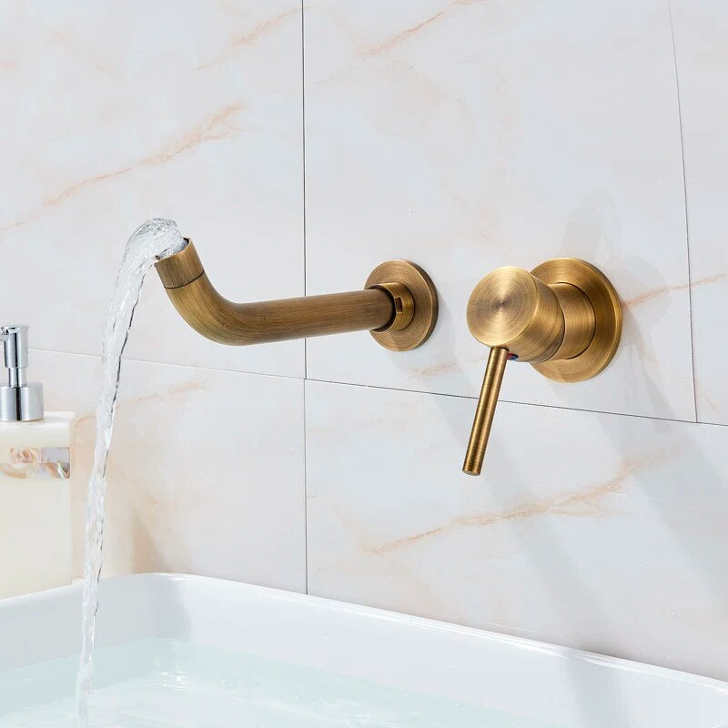 The Rozin Antique Brass Wall Mounted Bathroom Faucet pours water into a deep white sink, set against beige marble tiles. The steady, clear stream and minimalist design impart a modern aesthetic to the setup.