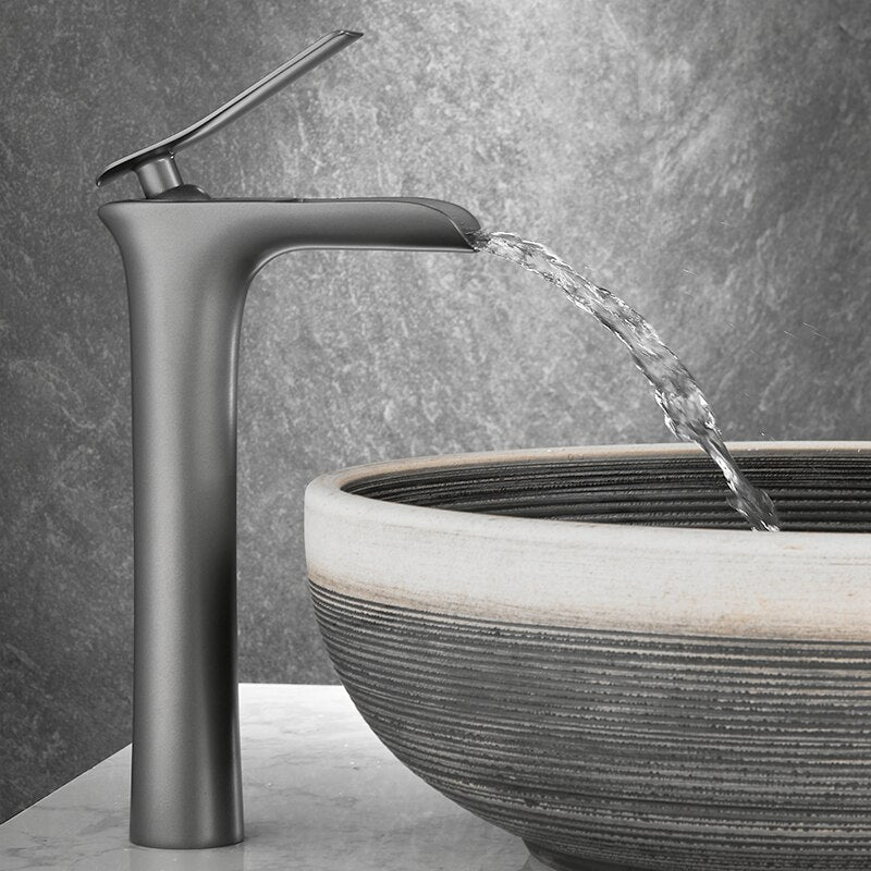 The Tiqui™ Gunmetal Single Handle Bathroom Sink Faucet, a tall and sleek stainless steel fixture with a modern design, dispenses water into a round, stylish stone sink set on a marble countertop. The background features a textured gray wall.