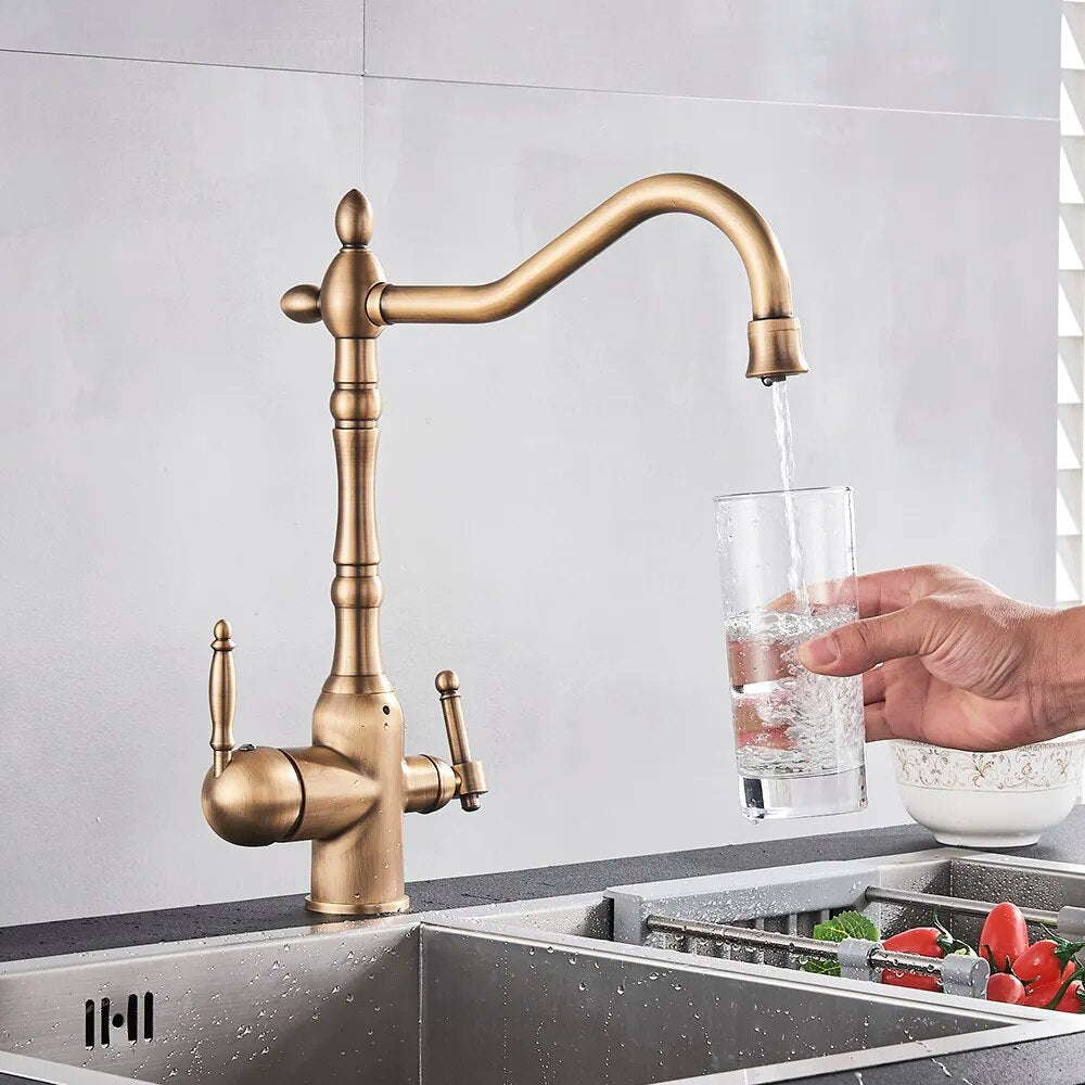 A sleek Uythner Dual Handle Solid Brass Faucet With Filtered Water Tap, featuring a vintage design, is mounted on a gray countertop. Water flows from the curved spout into a stainless steel sink below. In the background, a white bowl with a decorative pattern rests beside vegetables. Light filters through blinds, highlighting the elegant fixture.