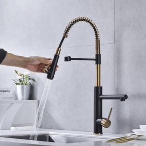 A modern kitchen sink with the stylish black and gold Tiqui™ Single Handle Pull Out Spout Kitchen Faucet by Smesiteli is seen. Water is flowing from the faucet into the sink. On the counter, there are white bowls, decorative items, a book titled "Company," and a potted plant.