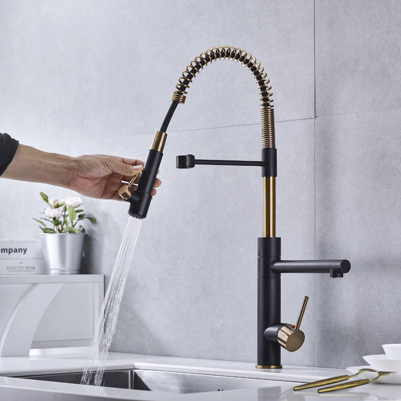 A modern kitchen sink with the stylish black and gold Tiqui™ Single Handle Pull Out Spout Kitchen Faucet by Smesiteli is seen. Water is flowing from the faucet into the sink. On the counter, there are white bowls, decorative items, a book titled "Company," and a potted plant.