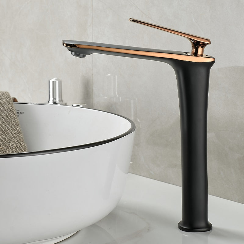 The Becola™ Single Handle Bathroom Faucet, with its modern matte black and rose gold finish, stands next to a white ceramic basin sink featuring a metallic rim. The faucet's sleek design includes a long, high arch and a lever handle. The background showcases light grey tiled walls.