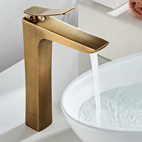 An Antique Bronze Deck Mounted Bathroom Sink Faucet by Tiqui is shown with water flowing from its spout into a white, round basin sink. The sleek design and minimalist aesthetics suggest a contemporary bathroom setting. The faucet handle is positioned upright.