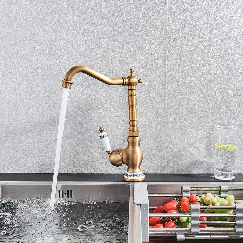 An Ellen Single Handle Antique Brass Kitchen Faucet, featuring a vintage-style design with a white ceramic handle and blue floral accents, is mounted on a marble countertop next to a sink filled with tomatoes and green peppers. A glass of water with lemon slices completes the charming setting.