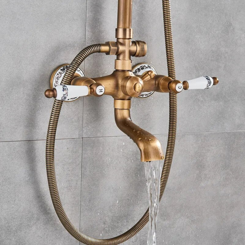 A wall-mounted Rozin Antique Brass Complete Shower Faucet Set with dual handles and white ceramic accents, labeled 'H' for hot and 'C' for cold. Water flows from the spout into a sink below, with flexible metal hoses connected to the faucet. The background is a gray tiled wall.