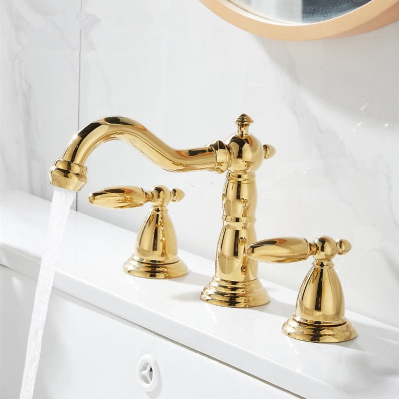 A Tiqui™ Solid Brass Contemporary Bathroom Sink Faucet, featuring a curved spout and two matching handles, is elegantly installed on a white marble countertop with a gray marble backsplash. Water flows from the faucet into the white sink below. A blue towel is partially visible on the left side.