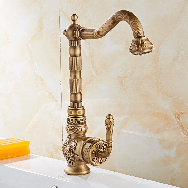 A Miho Single Handle Antique Bronze Faucet with intricate vintage-style designs is mounted on a marble countertop. The background features a beige marble-patterned wall, and a yellow soap bar is placed next to the faucet.