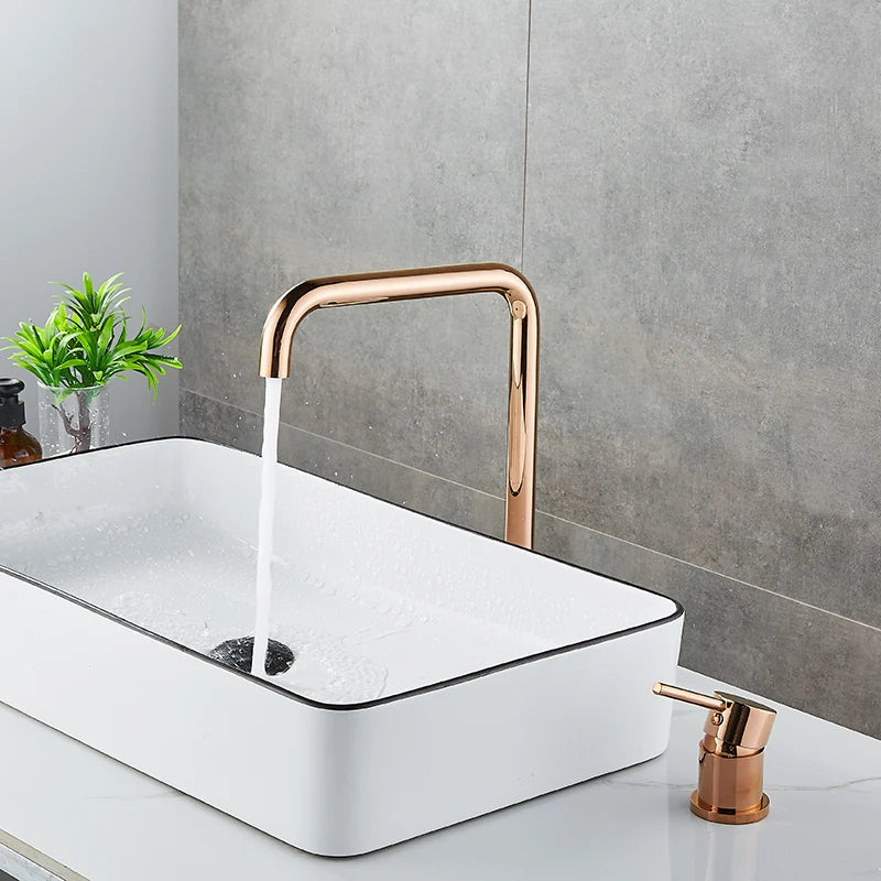 A modern bathroom sink setup featuring a rectangular white vessel sink with water flowing from AllFixture's sleek, curved Single Handle Rose Gold Bathroom Sink Faucet. Next to it is a matching rose gold soap dispenser. Complementing the scene is a grey tiled wall and a small plant adding a touch of green, reminiscent of chic kitchen faucet designs.