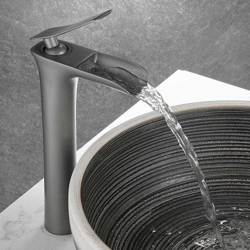 The Tiqui™ Gunmetal Single Handle Bathroom Sink Faucet, a tall and sleek stainless steel fixture with a modern design, dispenses water into a round, stylish stone sink set on a marble countertop. The background features a textured gray wall.