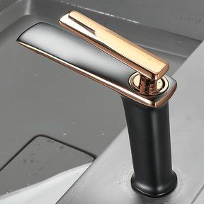 The Becola™ Single Handle Bathroom Faucet, with its modern matte black and rose gold finish, stands next to a white ceramic basin sink featuring a metallic rim. The faucet's sleek design includes a long, high arch and a lever handle. The background showcases light grey tiled walls.