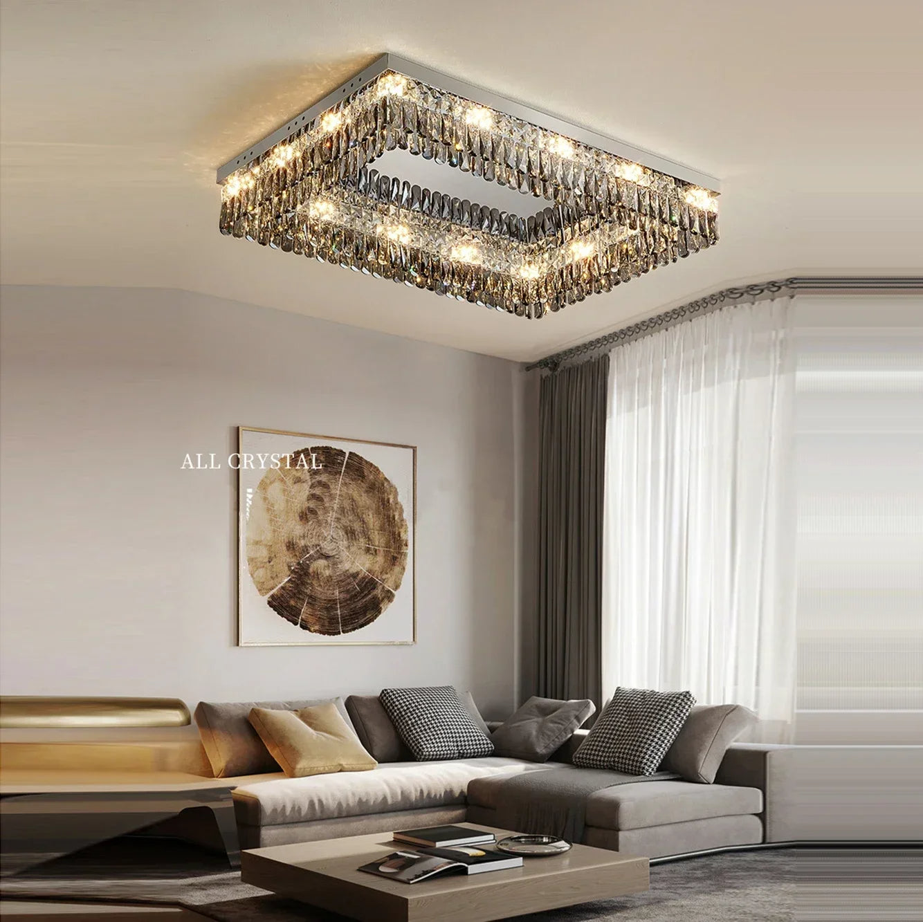 A modern living room with a handmade **Giano Crystal Ceiling Light by Morsale.com** hanging from the ceiling. The room features a gray sectional sofa adorned with throw pillows, a light brown coffee table with decor items, and a framed abstract art piece on the wall. Sheer curtains cover the window.