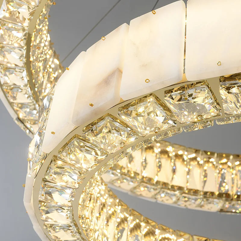 Close-up of a luxurious, modern ceiling chandelier featuring two interlocking, circular light fixtures. The lights are adorned with square-cut crystal accents and white marble-like panels, creating an elegant design. This Morsale.com Natural Marble & Crystal Modern Ceiling Light Fixture emits a warm, radiant glow.