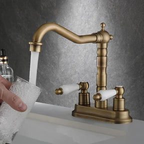 The "Antique Brass Bathroom Faucet - Timeless Elegance for Your Sink" by Allfixture stands over the sink, water flowing gracefully from its dual handle design with white handles. The background features a dark, marble-like surface. A soap dispenser is visible in the left corner of the image, complementing the premium brass construction.