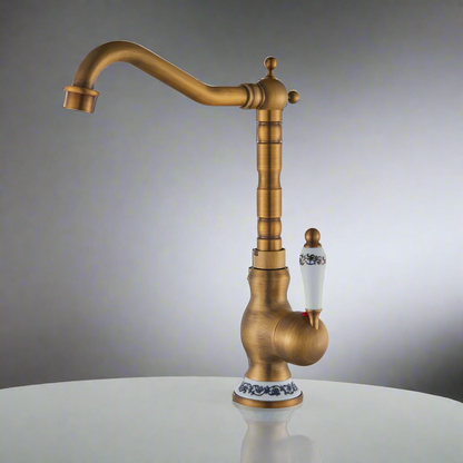 The Ellen Single Handle Antique Brass Kitchen Faucet features a vintage style with a high curved spout and a white porcelain handle with blue floral accents, crafted from solid brass in an antique bronze finish.