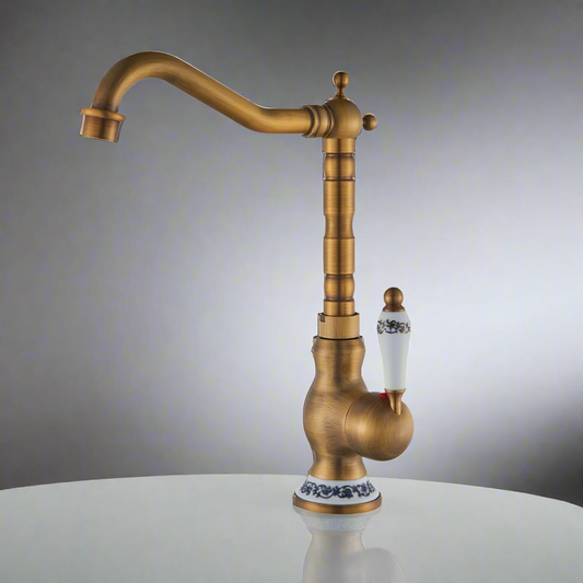 The Ellen Single Handle Antique Brass Kitchen Faucet features a vintage style with a high curved spout and a white porcelain handle with blue floral accents, crafted from solid brass in an antique bronze finish.