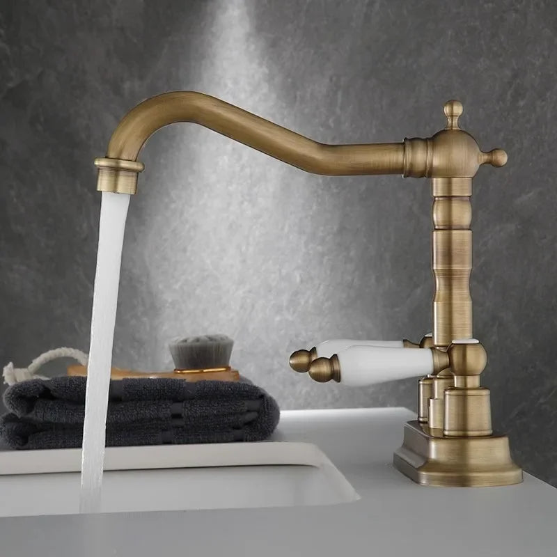 The "Antique Brass Bathroom Faucet - Timeless Elegance for Your Sink" by Allfixture stands over the sink, water flowing gracefully from its dual handle design with white handles. The background features a dark, marble-like surface. A soap dispenser is visible in the left corner of the image, complementing the premium brass construction.