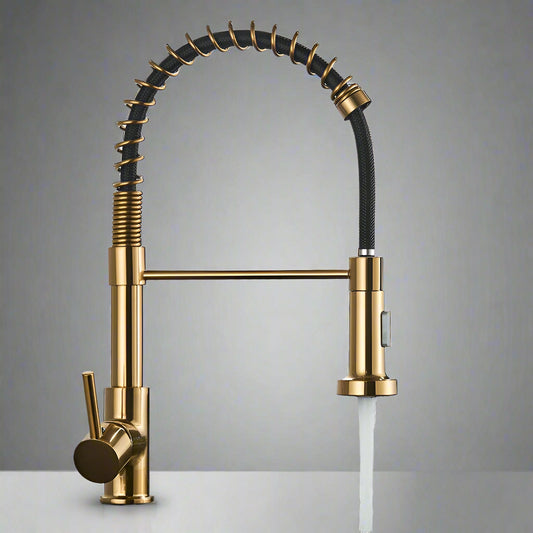 The Quyanre Pro Spring Neck Pull-Down Kitchen Faucet, in a modern gold finish with a flexible coiled neck and single lever handle, is mounted on a white countertop. Water streams from its pull-down spray head against a plain gray wall.