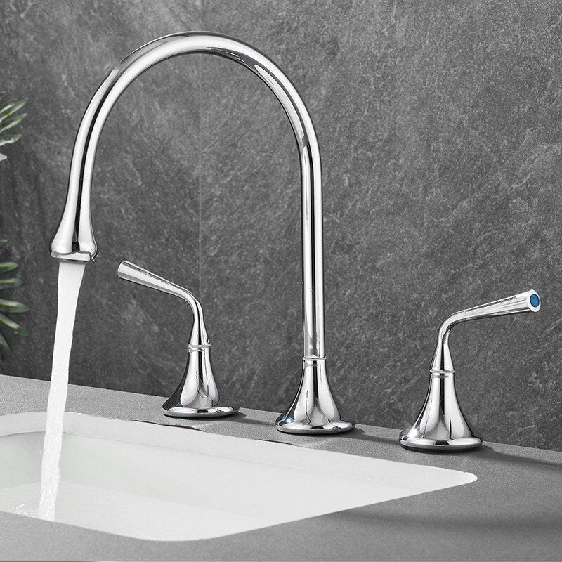 A sleek Tiqui™ Solid Brass Double Handle Bathroom Faucet by Mike Jake, featuring a high-arc spout, is mounted on a grey countertop. Water flows gracefully from the spout into a white sink. The background showcases a grey, textured wall and some green plant leaves peeking in from the left edge.