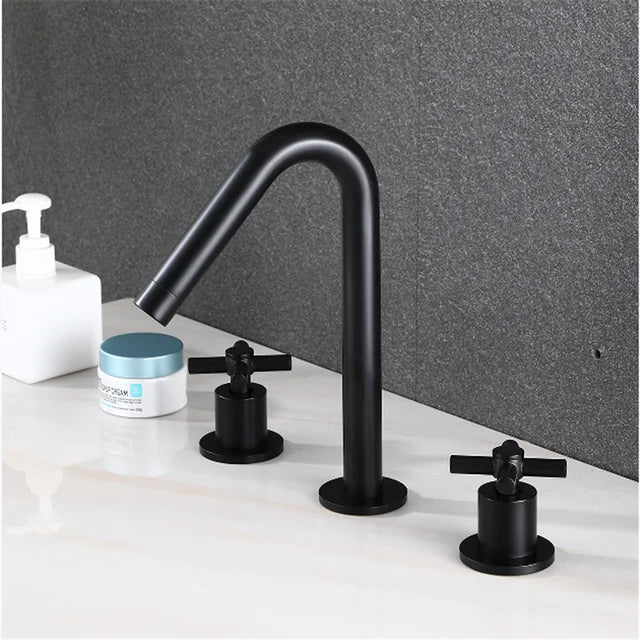 A modern bathroom sink features the Allfixture Basin Faucet - 3 Hole Widespread Bathroom Mixer Tap with a luxurious gold finish and two matching black handles. A white liquid soap dispenser and a small blue and white container are placed on the countertop, which is set against a dark textured wall.