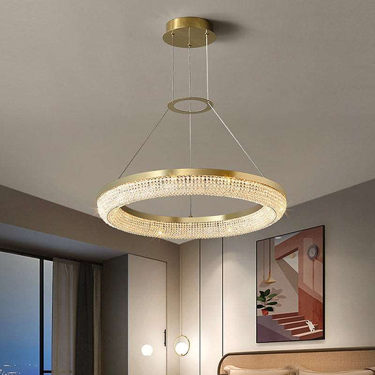 A modern interior showcases the Bergamo Ceiling Light Fixture from Morsale.com—a circular, gold LED chandelier adorned with crystal accents—suspended elegantly from the ceiling. Below it rests a bed with a tufted headboard, accompanied by an art piece adorning the wall beside it. The room is further enhanced by a large window and ambient wall lighting.