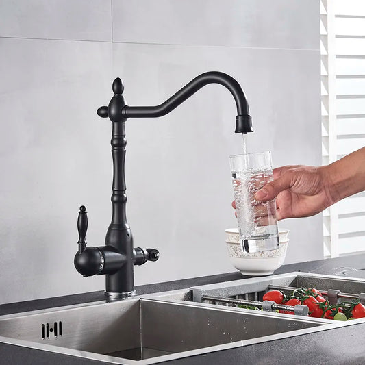 A hand fills a glass with water from the Uythner Heritage Kitchen Faucet in black. Below, a strainer holds washed tomatoes and vegetables, set against a modern tiled wall.
