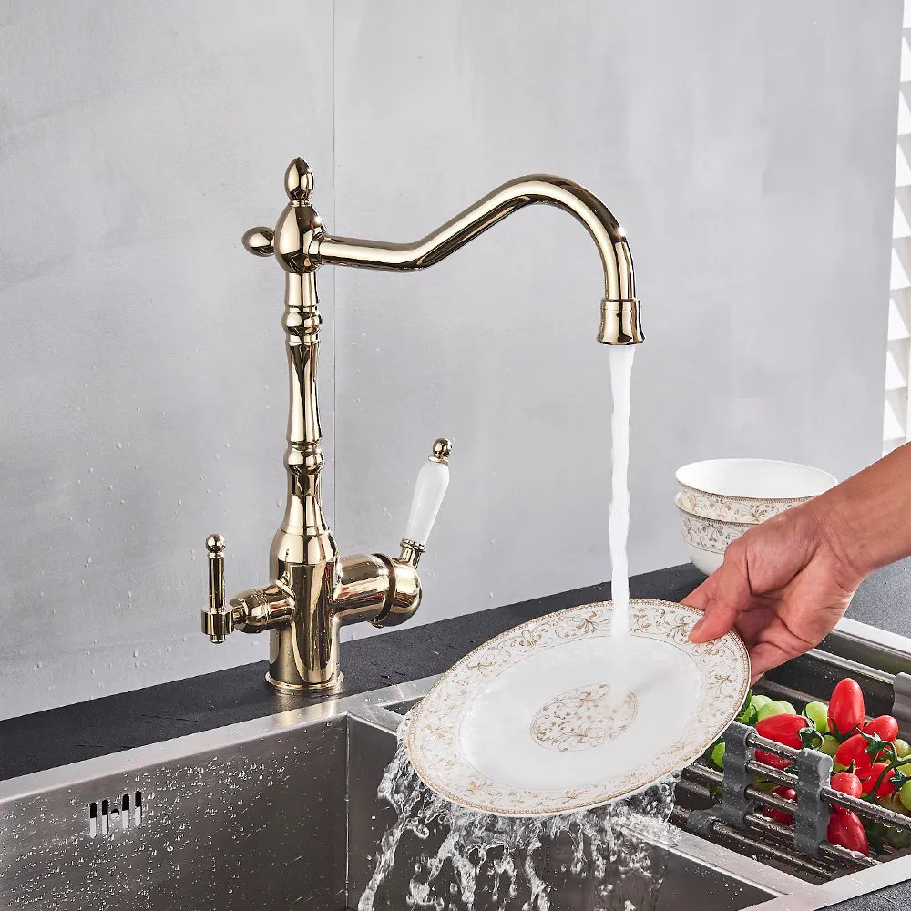 A polished gold kitchen faucet with a filtered water tap and vintage design by Uythner is mounted on a countertop. Water is flowing from the faucet into a clear glass being held by a person's hand. A small dish and green herbs are visible in the background.