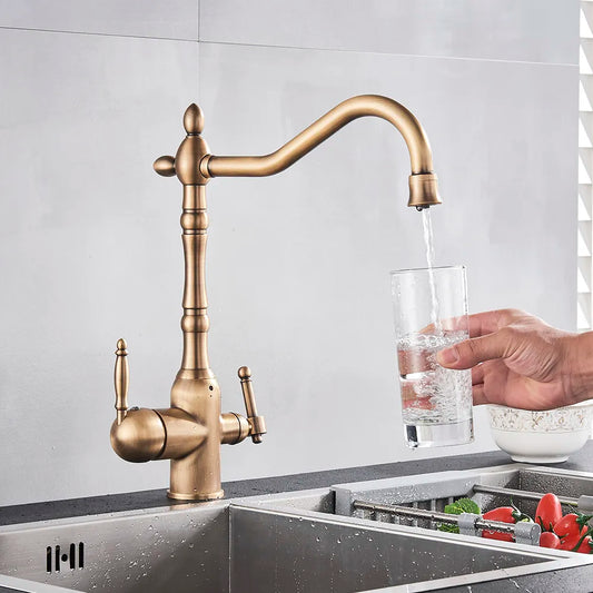 A hand holds a glass under the Uythner Heritage Kitchen Faucet in Antique Brass as water flows. The warm kitchen scene, with vegetables and produce nearby, highlights the vintage charm of this elegant fixture.
